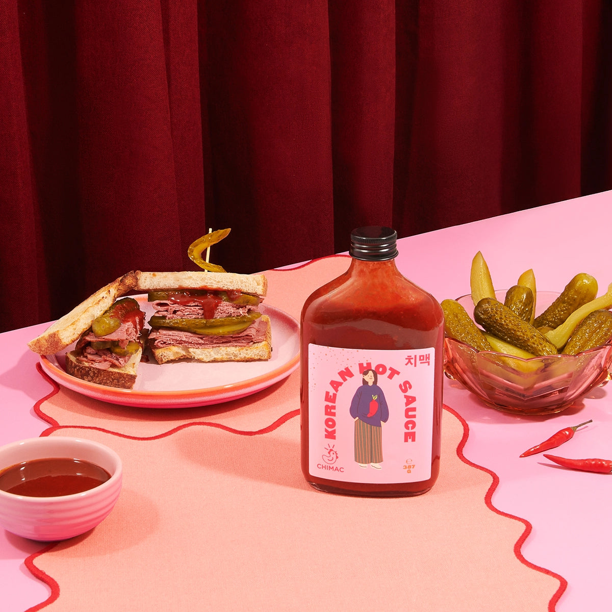 Korean hot sauce bottle with a sandwich and pickles on a pink tablecloth.